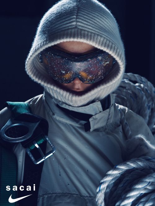 Person in hooded jacket and goggles holds coiled rope, Sacai and Nike logos visible in the foreground against dark background.