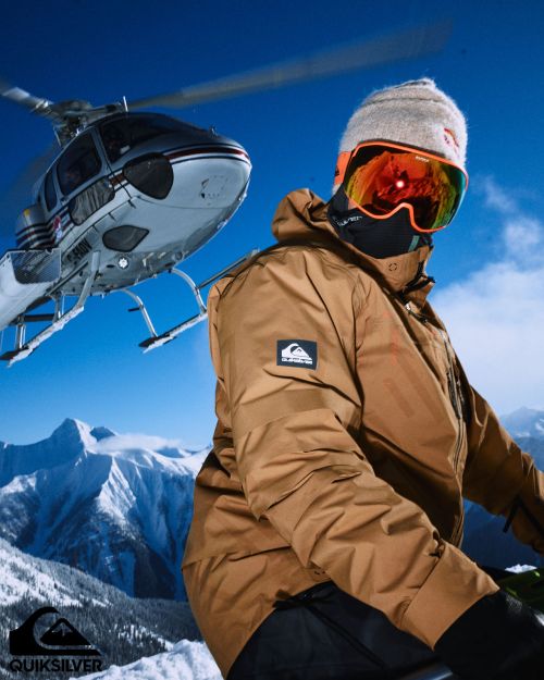 Man in ski gear with goggles and a beanie in snowy mountain landscape, helicopter in background, Quiksilver logo at the bottom left.