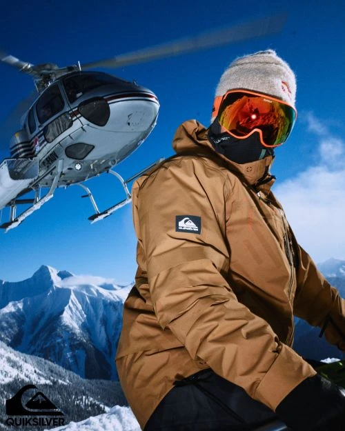 Man in ski gear with goggles and a beanie in snowy mountain landscape, helicopter in background, Quiksilver logo at the bottom left.