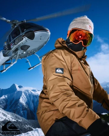 Man in ski gear with goggles and a beanie in snowy mountain landscape, helicopter in background, Quiksilver logo at the bottom left.