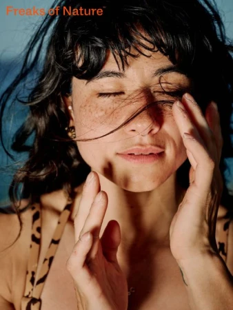 Close-up portrait of a woman with freckles and dark hair holding her hands to her face outdoors against a blue sky.