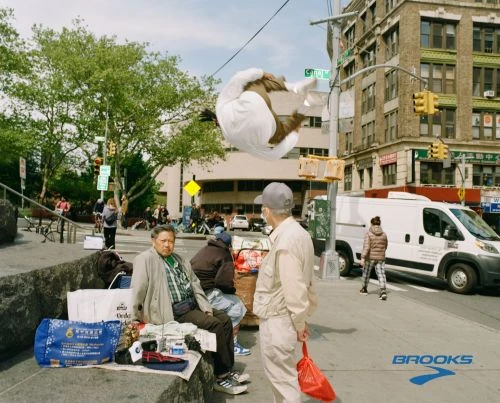 Street scene shows a man in mid-air performing a flip, with other people, a building, and trees in the background on a sunny day.