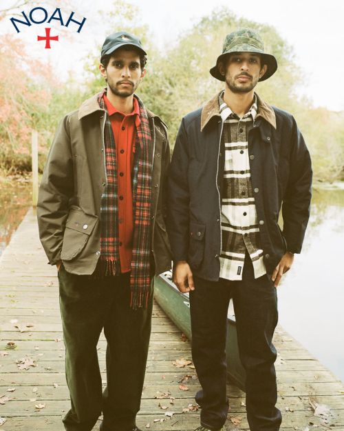 Two men pose on a wooden dock in outerwear and hats near water and foliage in a medium outdoor shot. The Noah logo is visible.
