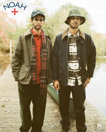 Two men pose on a wooden dock in outerwear and hats near water and foliage in a medium outdoor shot. The Noah logo is visible.