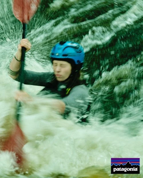 An adult female kayaking in whitewater rapids wearing a blue helmet, holding a red paddle, surrounded by splashing water in an outdoor setting.