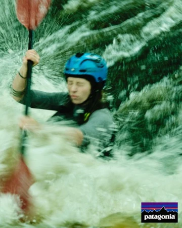 An adult female kayaking in whitewater rapids wearing a blue helmet, holding a red paddle, surrounded by splashing water in an outdoor setting.