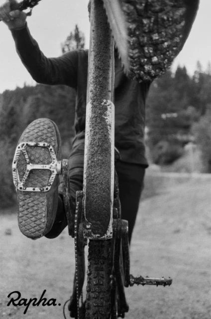 Monochrome shot of a cyclist on a muddy bike path with a foot on the pedal and trees blurred in the background.