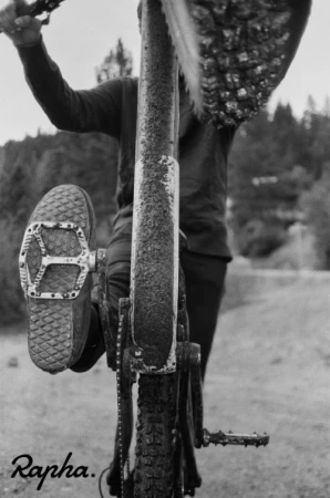 Monochrome shot of a cyclist on a muddy bike path with a foot on the pedal and trees blurred in the background.