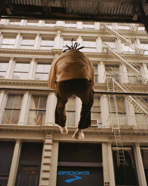Low angle view of a person wearing a tan sweater and dark pants hanging from a building with fire escapes and a Brooks logo.