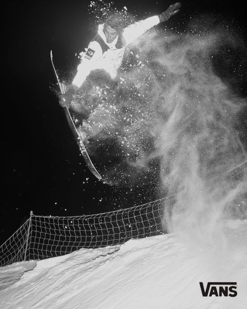 A snowboarder in a white snowsuit jumps high above the snow, kicking up a cloud of powder against a dark, wintry background in a black and white action shot.