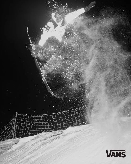 A snowboarder in a white snowsuit jumps high above the snow, kicking up a cloud of powder against a dark, wintry background in a black and white action shot.