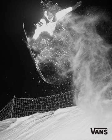 A snowboarder in a white snowsuit jumps high above the snow, kicking up a cloud of powder against a dark, wintry background in a black and white action shot.