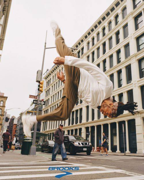 An acrobatic adult male is doing a backflip on a crosswalk in an urban setting with buildings and street signs visible in the background.