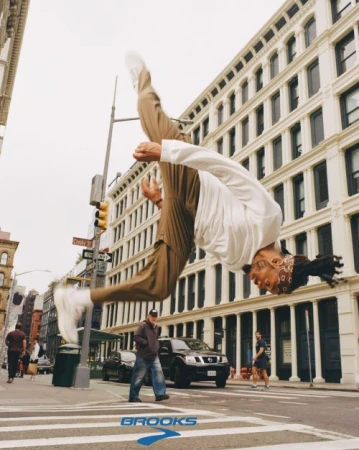 An acrobatic adult male is doing a backflip on a crosswalk in an urban setting with buildings and street signs visible in the background.