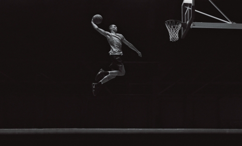Monochromatic image of a basketball player in mid-air with a raised arm, holding a ball near a basketball hoop and net in a dark indoor setting.