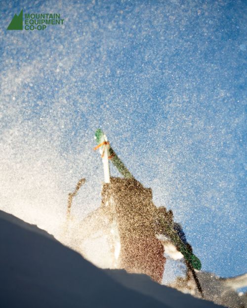 Action shot of a snowboarder with snow spraying in the air against a partly cloudy blue sky, capturing the dynamic movement of the sport.