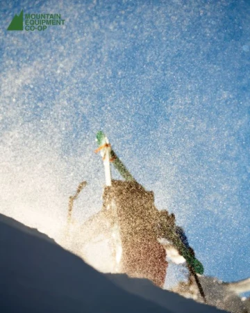 Action shot of a snowboarder with snow spraying in the air against a partly cloudy blue sky, capturing the dynamic movement of the sport.