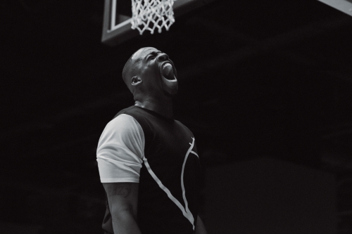 Athletic African-American man in sportswear screams in triumph by a basketball hoop with a black background in monochrome photography.
