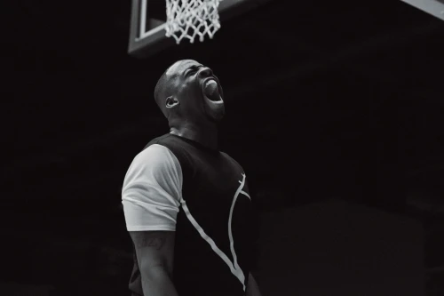 Athletic African-American man in sportswear screams in triumph by a basketball hoop with a black background in monochrome photography.