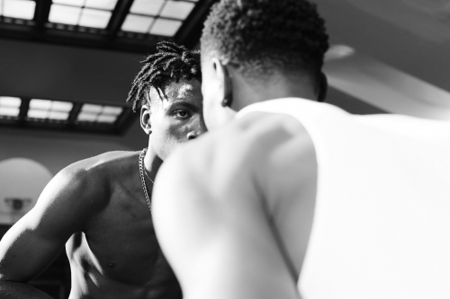Two Black adult men face each other indoors, one shirtless and wearing a chain, with the other wearing a tank top in black and white.