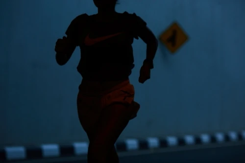 Silhouette of an adult female running in the dark, wearing sports clothing, with a road sign and striped curb in the background.