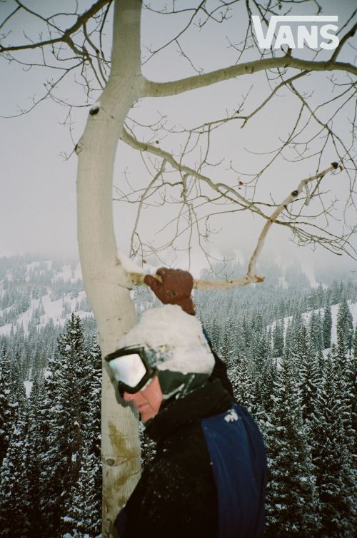 A person in ski goggles and winter clothing stands in a snowy forest next to a snow-covered aspen tree on a foggy day.
