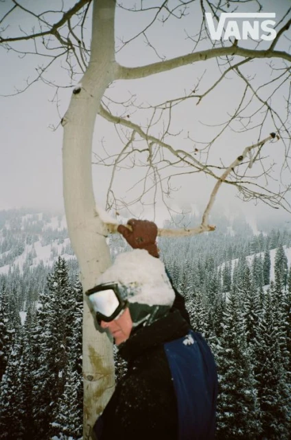 A person in ski goggles and winter clothing stands in a snowy forest next to a snow-covered aspen tree on a foggy day.