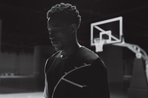 Black and white portrait of an adult male basketball player in a gym with a blurred basketball hoop in the background.
