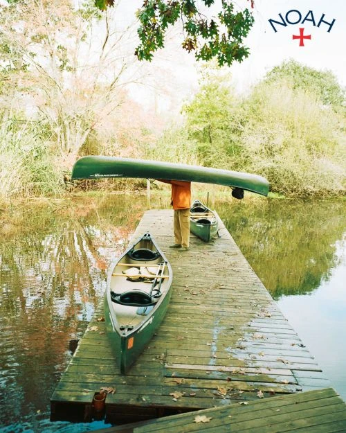 Person carries green canoe overhead on a wooden dock near water with two more canoes; trees in the background, outdoors, daylight.