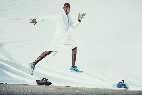 A man in a white robe jumps for joy in blue running shoes against a white background, with bags on a grey surface in the foreground.