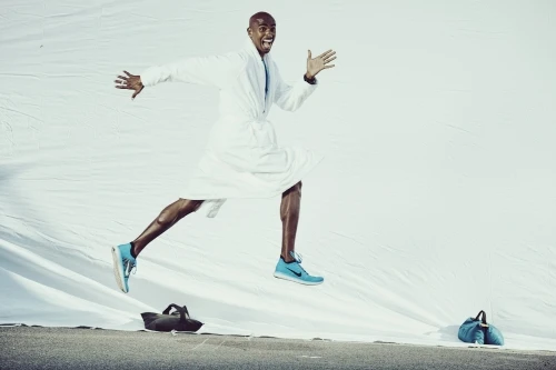 A man in a white robe jumps for joy in blue running shoes against a white background, with bags on a grey surface in the foreground.