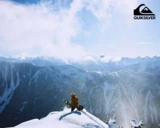 Snowboarder standing on a snowy mountain ridge holding a snowboard under an expansive blue sky with distant peaks.