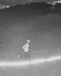 Monochrome action photograph by Cole Barash of a runner moving across a grassy slope illuminated by a bright horizontal light trail.