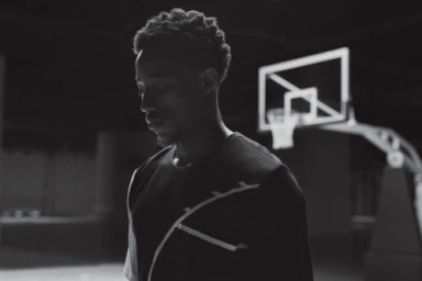 Black and white portrait of a black man wearing a dark t-shirt with a basketball hoop in the dim background by Cole Barash