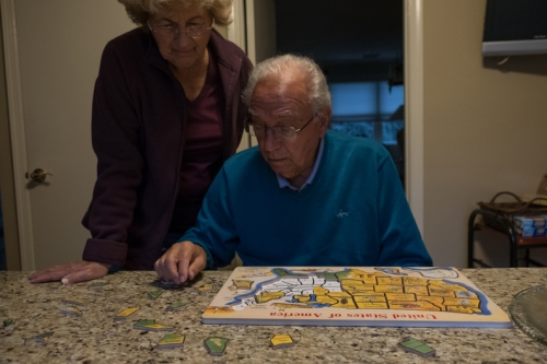 An elderly couple works together on a jigsaw puzzle of the United States at a table in a home, illuminated by indoor lighting.
