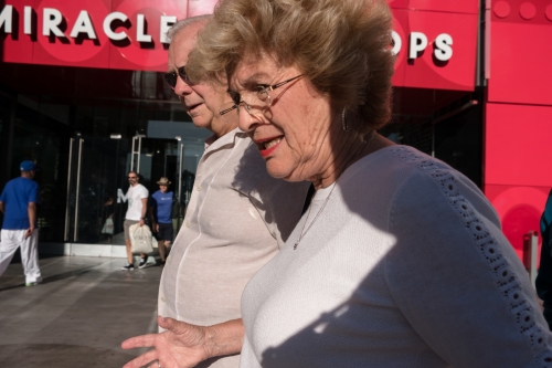 Two elderly people walk in an urban environment with a red building and other people in the background. The woman is smiling.