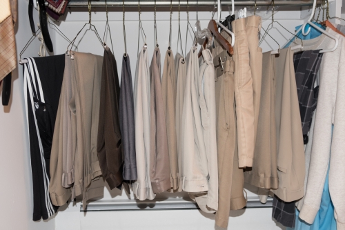 Several pairs of pants hang in a closet on a metal rod; the pants are in shades of beige, tan, gray, and black with white walls.
