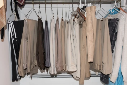Several pairs of pants hang in a closet on a metal rod; the pants are in shades of beige, tan, gray, and black with white walls.