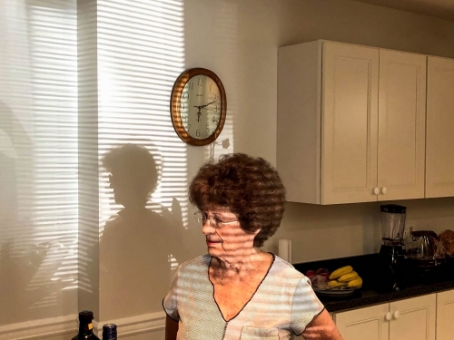 Mature woman in kitchen stands near countertop with food, shadowed by window blinds with wall clock and cabinets in the background.