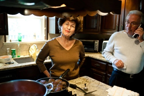 An older woman stands in a kitchen with a frying pan as an older man talks on a cell phone. Dark wood cabinets and granite counters.
