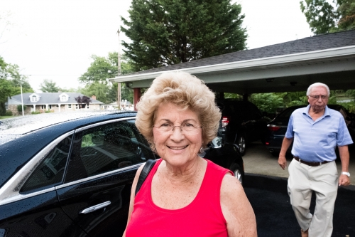 Elderly woman in red top smiles next to black car outside, with an elderly man walking behind a vehicle under the building's overhang.