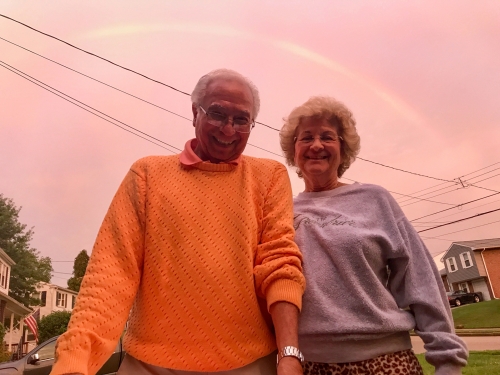 Two elderly people smile beneath a pink-hued sky, potentially at sunset or sunrise, a faint rainbow arc visible in the sky above.