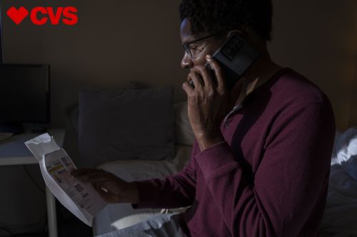 Adult Black man wearing glasses sits in bed at night, reviewing paperwork and talking on a cell phone in a dimly lit room.