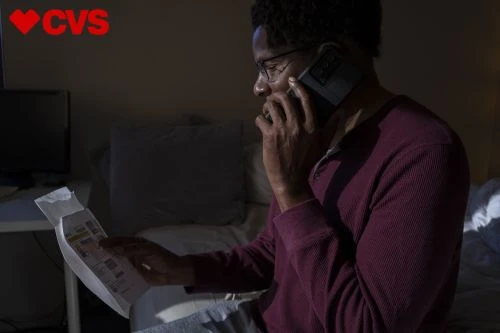 Adult Black man wearing glasses sits in bed at night, reviewing paperwork and talking on a cell phone in a dimly lit room.