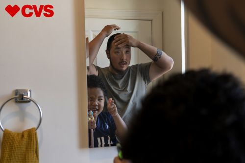 Father and son look in the mirror inside a bathroom featuring a towel and CVS logo, capturing a candid family moment.