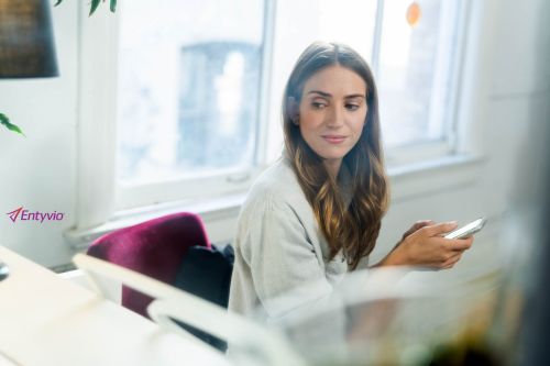 Woman looks at a cell phone near a bright window. She sits at a desk with a computer and plant visible.