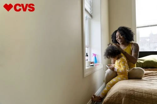 Mother brushing her child's hair next to a bright window in a bedroom. The child is seated on the bed and holding a smartphone.