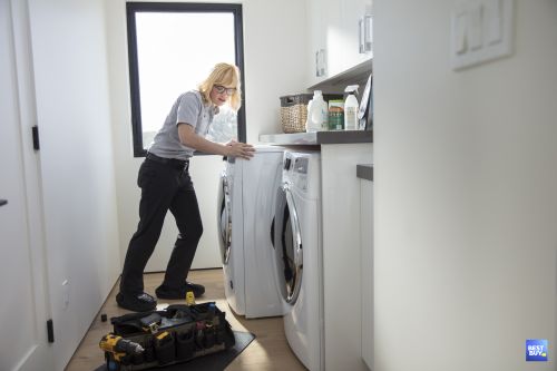 Adult female repair person leaning over a front-loading washing machine with a tool bag in a bright, indoor laundry room setting.