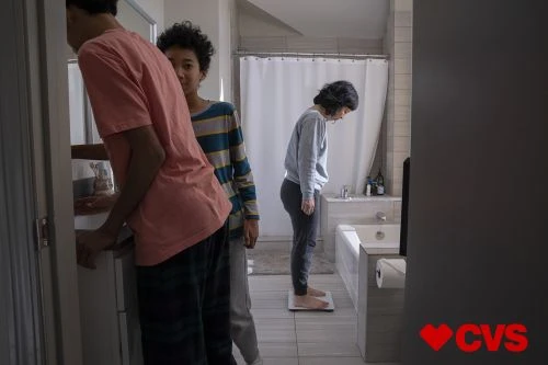 Woman stands on scale in bathroom, flanked by two boys near sink; promotes CVS brand with logo in corner, interior home setting.