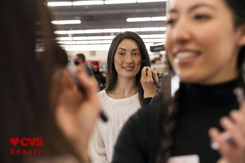 Two smiling women applying makeup in a retail store, with bright lighting and makeup tools and products visible on shelving units.